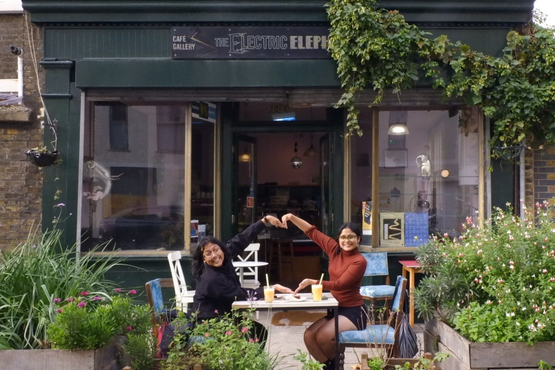 Two women are sat at a table in front of the cafe with fresh juice and food on it. They are smiling. There are planters and a plant climbing high 