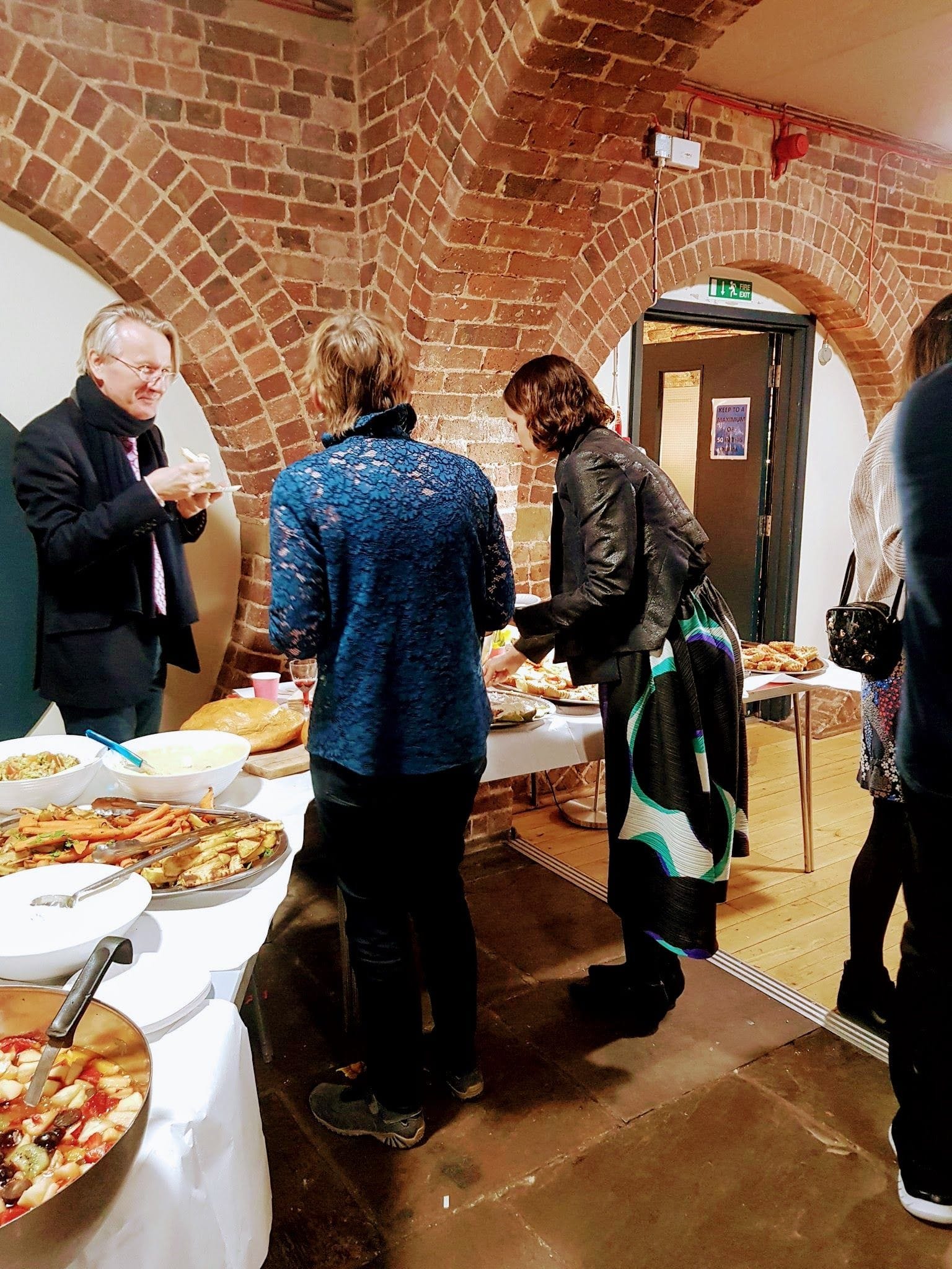 People helping themselves to food on large silver platters and a large bowl of fruit salad at a buffet 