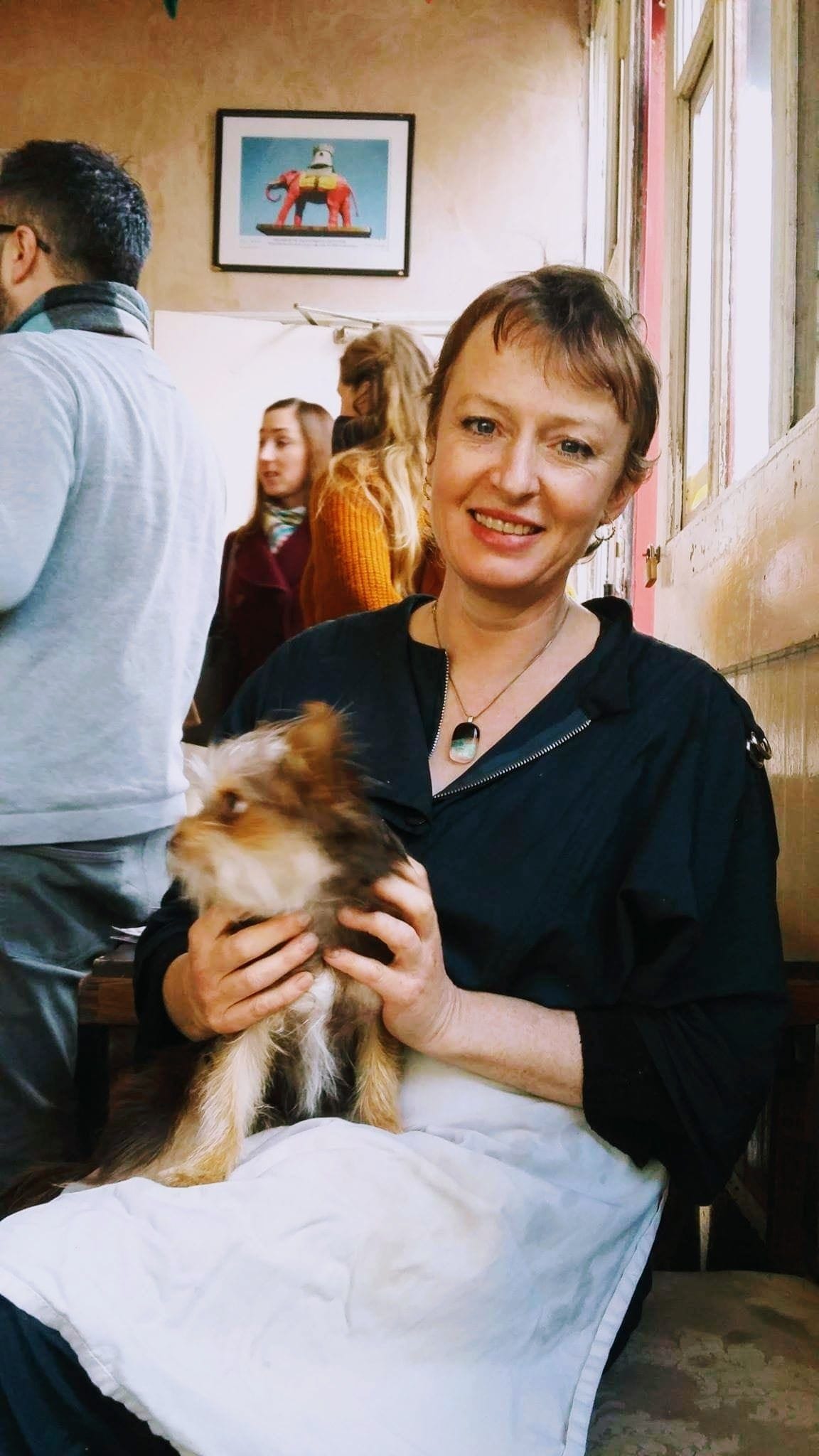 Louisa the cafe owner wearing a white apron, sat holding amall Yorkshire terrier, she is smiling andcustomers areat the counter behind her